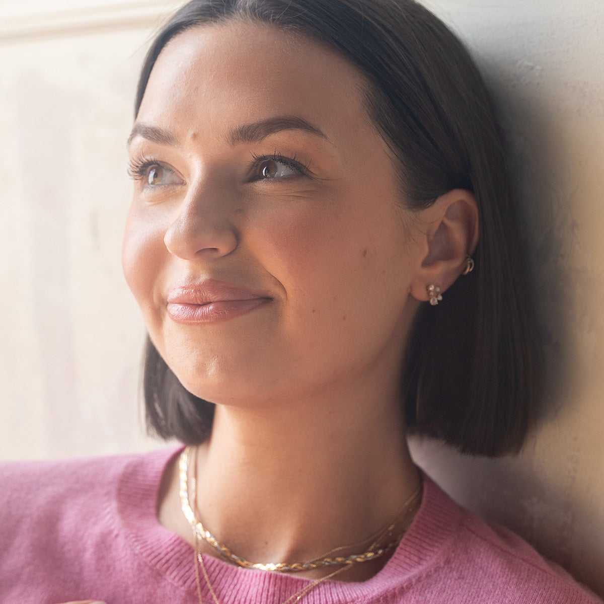 A woman with short dark hair and a pink top smiles softly while looking off to the side, wearing Solid Gold Soft Glow Pearl and Moonstone Drop Studs, paired with layered necklaces by a light-colored wall.
