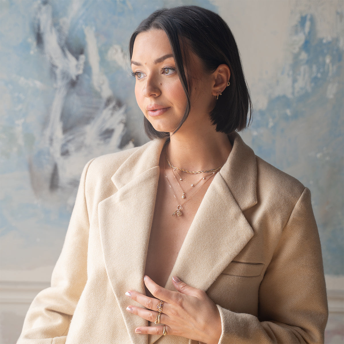 A woman with short dark hair, wearing a beige blazer and the Solid Gold Horizon Moonstone and Opal Sprinkle Necklace, stands indoors looking thoughtfully to the side against a blue and white abstract background.