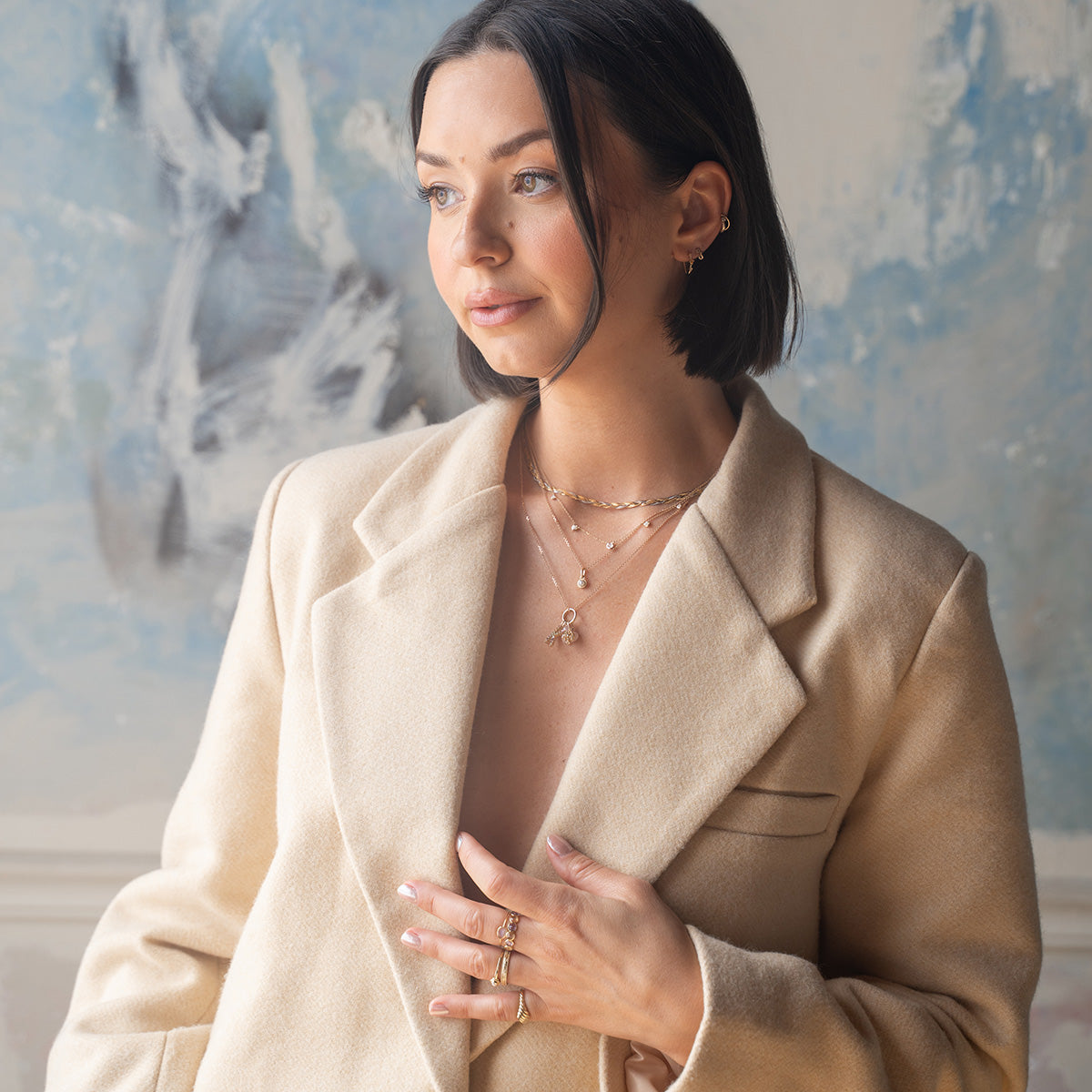 A woman with a short dark bob, cream-colored coat, and the Solid Gold Soft Glow Pearl Necklace stands against a blue and gray abstract background, looking to the side.
