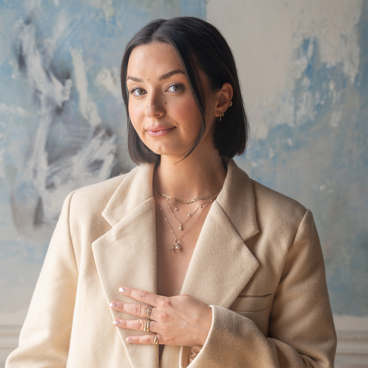 A woman with a bob haircut wears a cream coat and layered gold necklaces, standing against a blue and beige wall. She smiles gently, holding her hand to her chest to display the Solid Gold Sunrise Citrine and Alexandrite Band.
