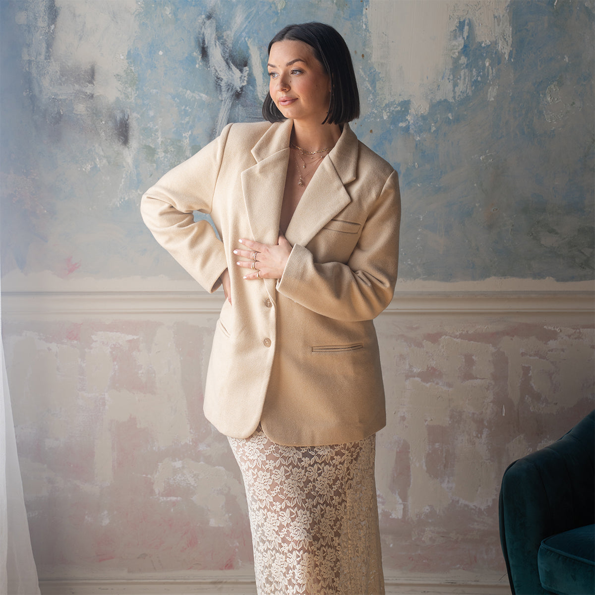 A woman with short dark hair stands indoors against a textured blue and beige wall, wearing a cream blazer, long white lace skirt, and the Solid Gold Pastel Gemstone Letter Charm, posing thoughtfully with one hand on her hip.