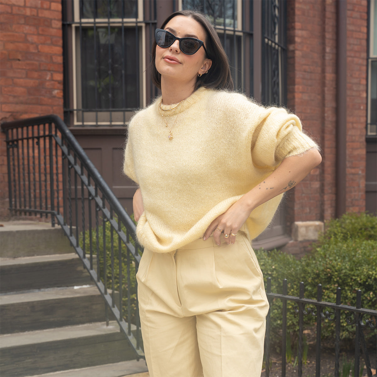 A woman in sunglasses, a yellow sweater, and matching high-waisted pants poses by a brick building and stairs, accessorized with the Solid Gold Horizon Moonstone and Opal Sprinkle Necklace—one hand in her pocket, the other on her hip.
