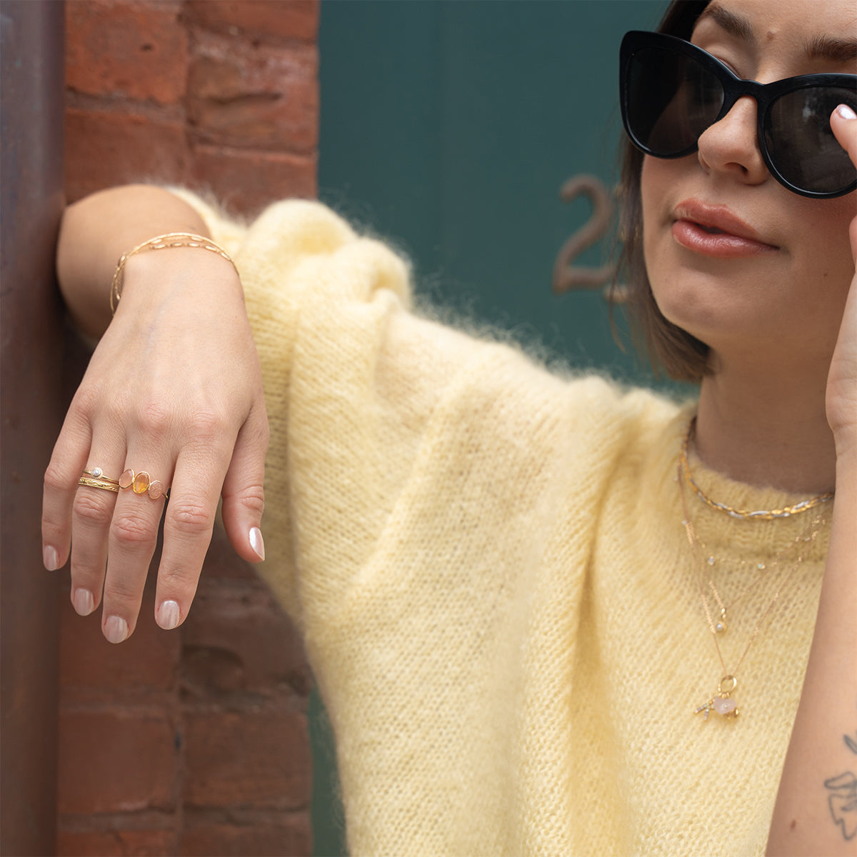 A woman in a soft yellow sweater leans against a brick wall, wearing the Solid Gold Morning Light Citrine and Sunstone Ring. She has a subtle forearm tattoo and adjusts her black sunglasses with one hand.