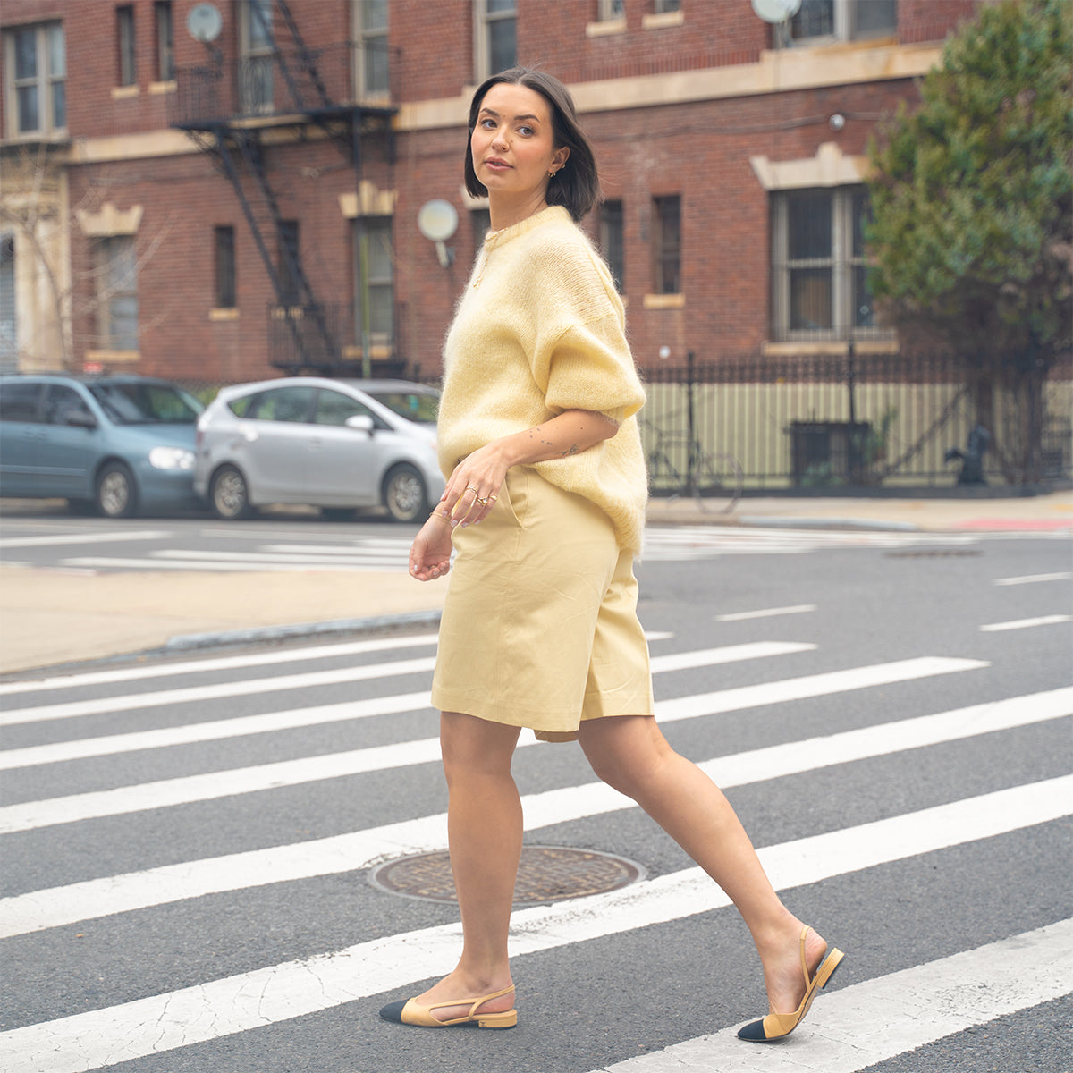 A woman walks across a city crosswalk on a cloudy day, wearing a fuzzy yellow sweater, beige shorts, yellow flats, and the Solid Gold Soft Glow Pearl Necklace. Brick buildings and parked cars are in the background.