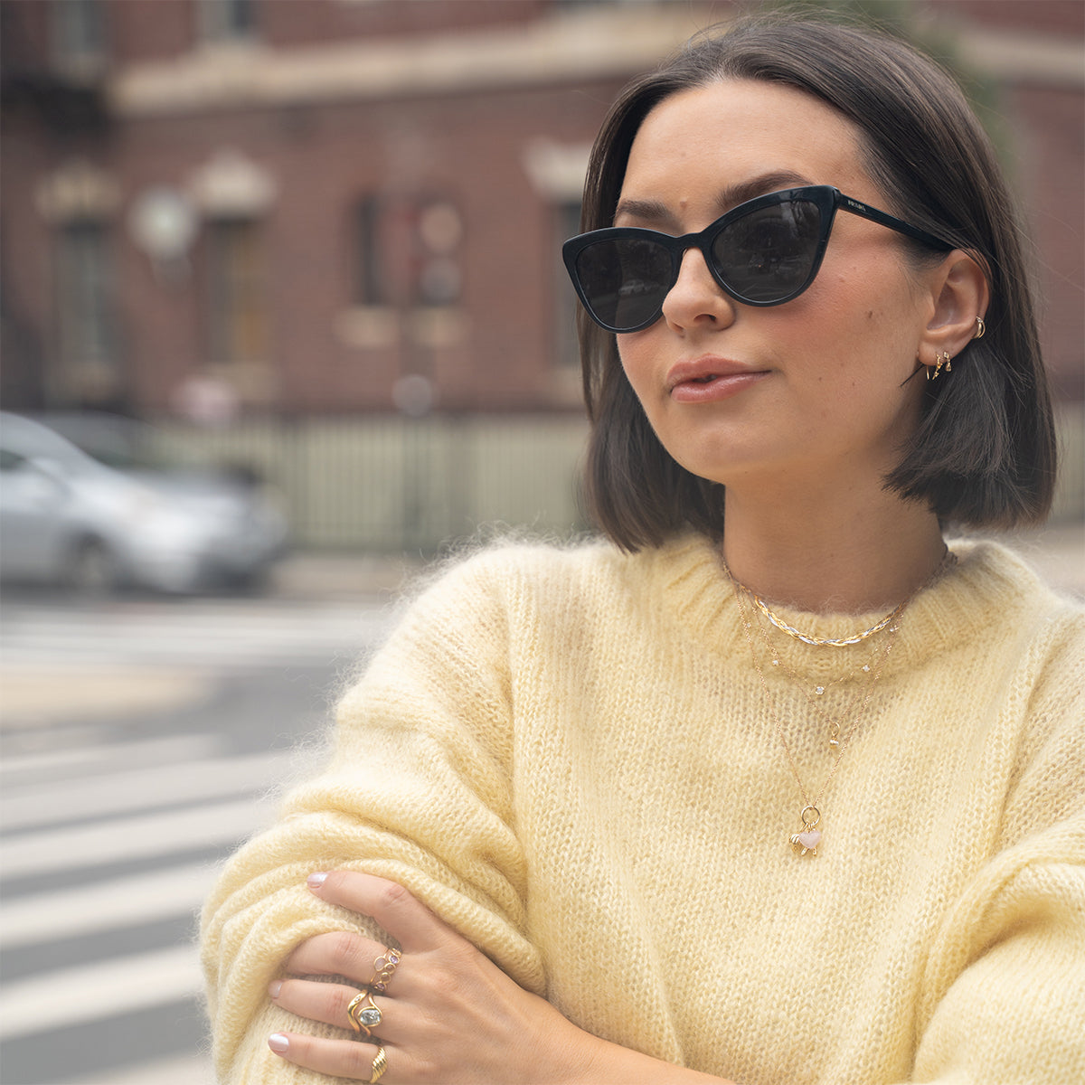 A woman with short dark hair, black cat-eye sunglasses, a pale yellow sweater, layered gold necklaces, and a Solid Gold Sunrise Citrine and Alexandrite Band stands confidently with arms crossed on a city street.