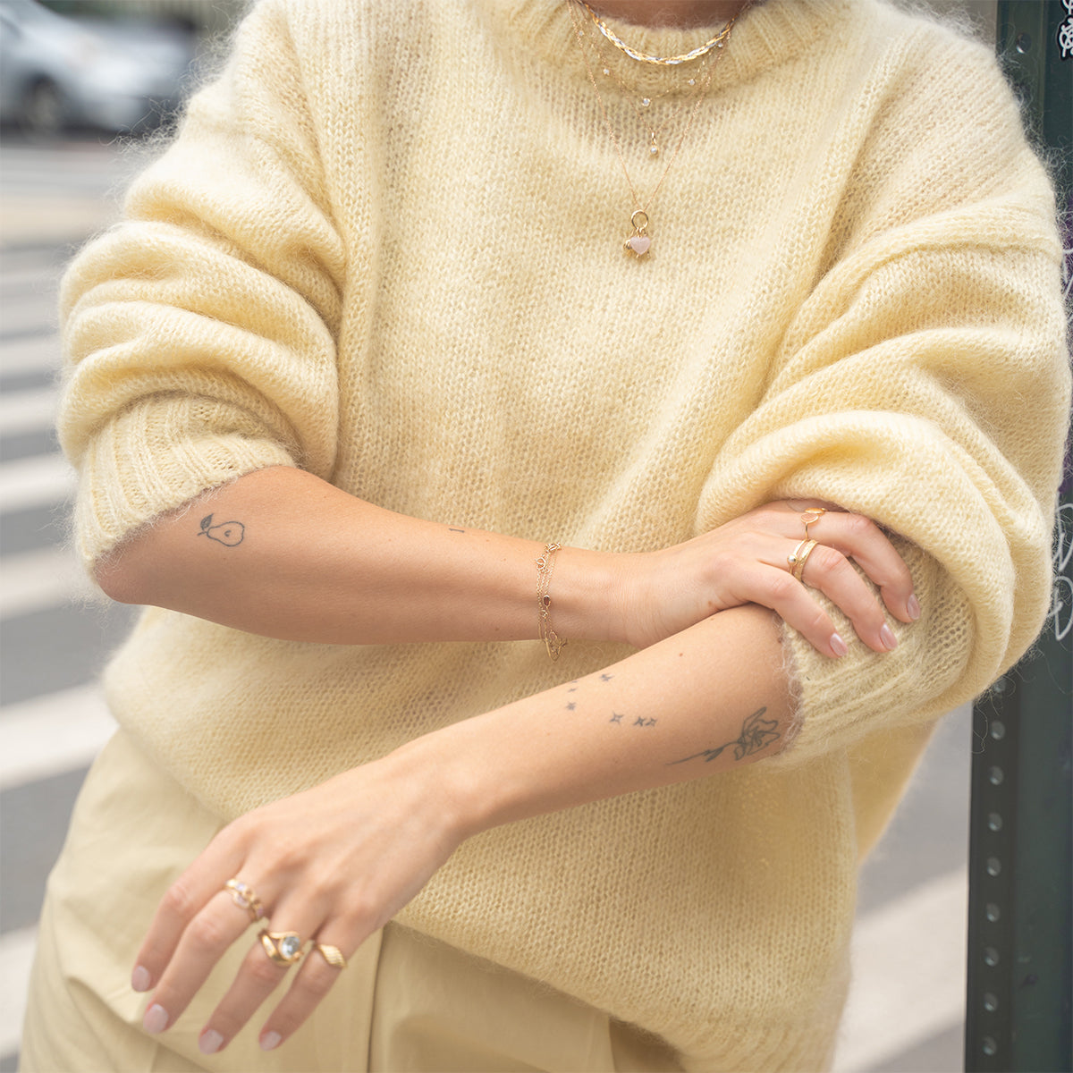 A person in a soft yellow sweater and matching pants wears gold rings, layered necklaces, and a Solid Gold Daybreak Moonstone and Blue Topaz Bracelet. Small star and fish tattoos decorate their forearms; the street scene behind them is blurred.