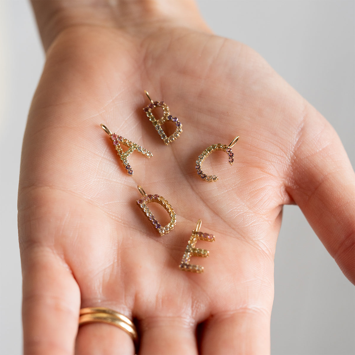 A hand holds four Solid Gold Pastel Gemstone Letter Charms shaped as A, B, C, and E. The person is also wearing two gold rings on their ring finger.