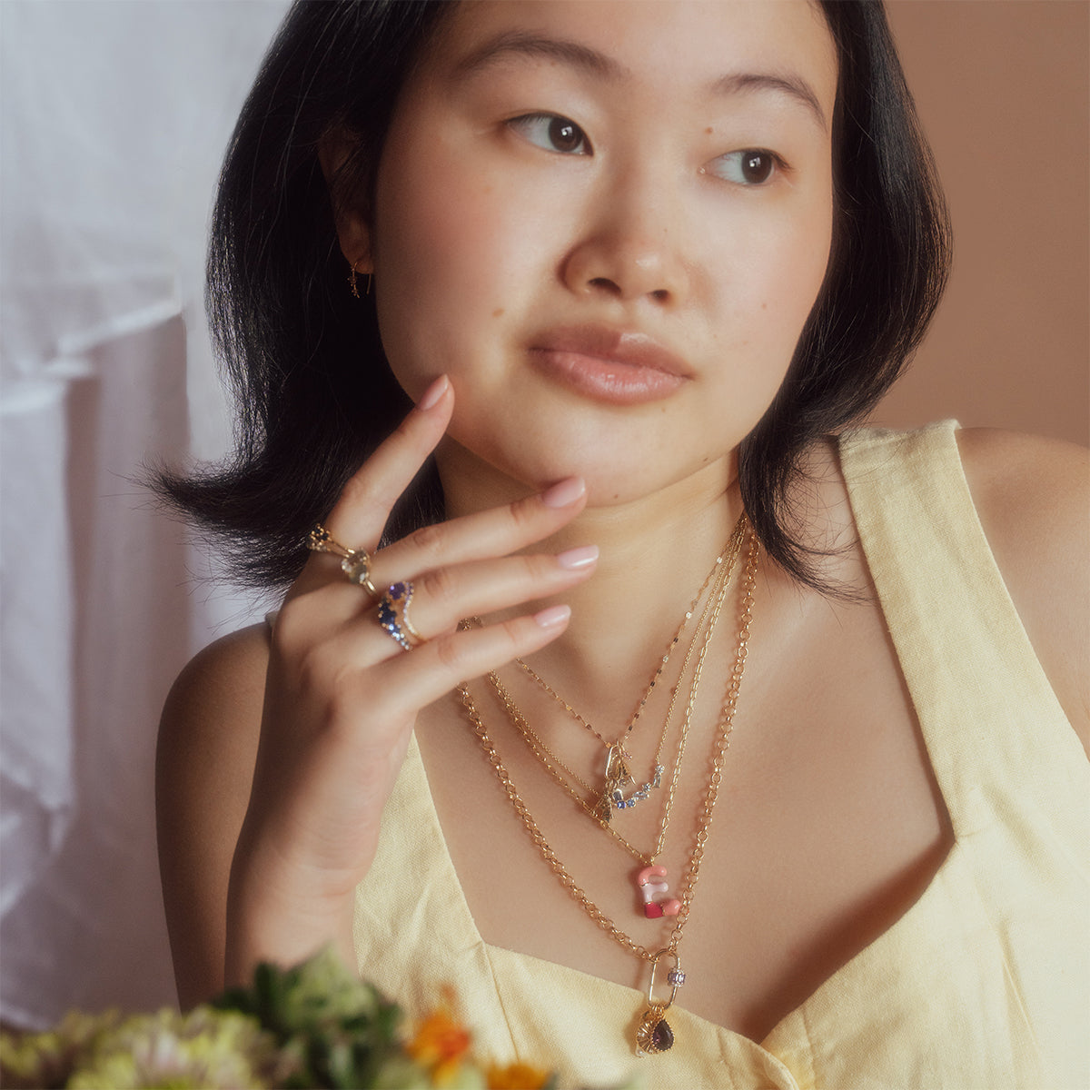 A woman in a yellow sleeveless top poses thoughtfully, chin on her hand. She wears layered rings and the Shades of You Ombre Birthstone Necklace, with soft natural lighting and blurred flowers in the foreground.
