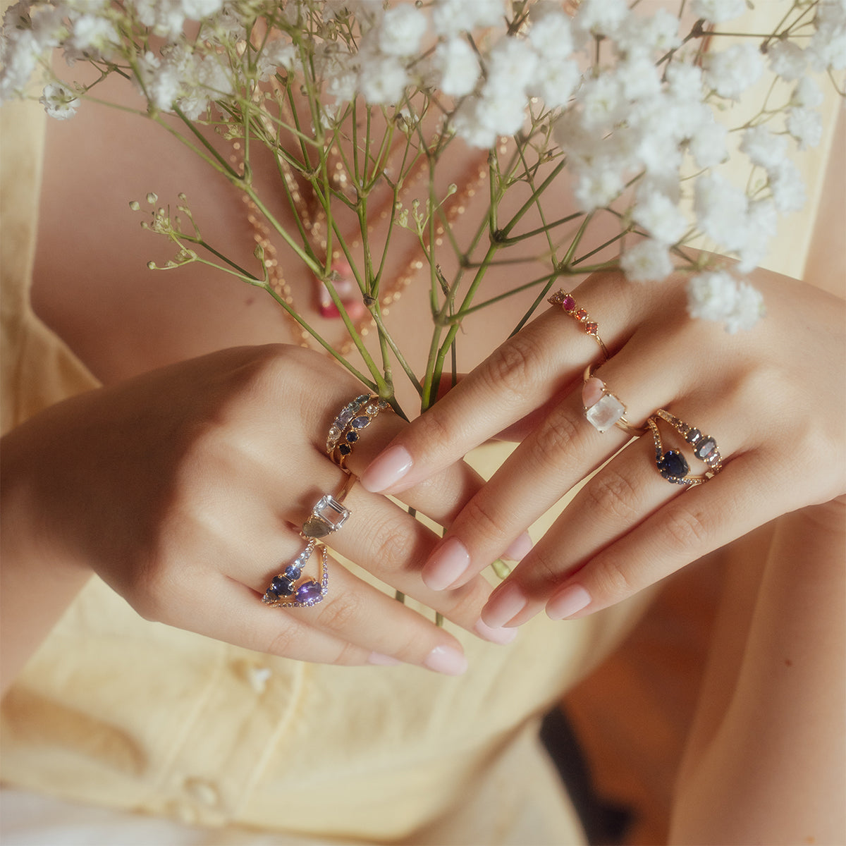 A woman in a pale yellow dress holds a bouquet of babys breath. Her manicured hands are adorned with gold rings, including the True Colors Classic Ombre Birthstone Ring and a dazzling 14k gold plated ring with blue gemstones.