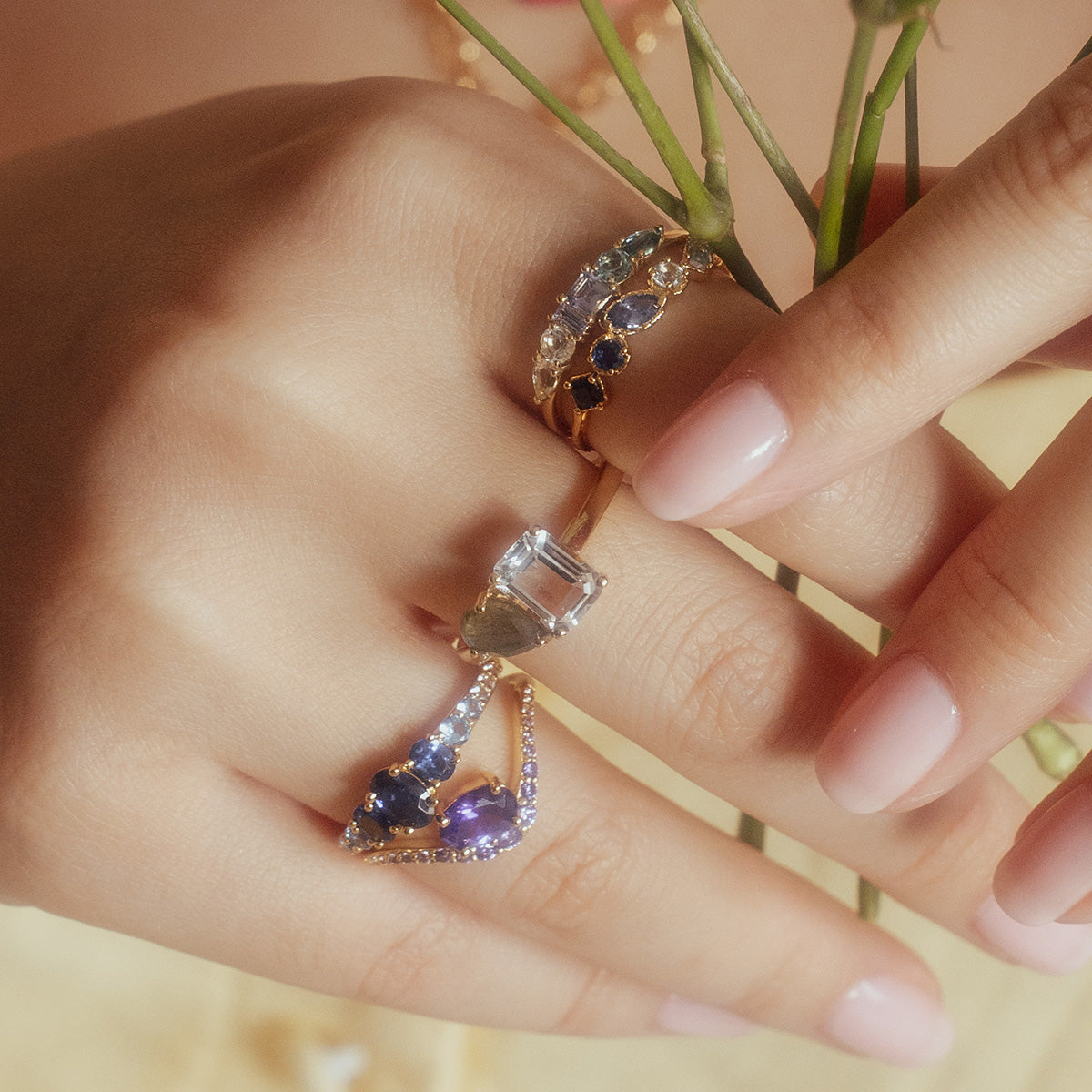A close-up of a hand with light pink manicured nails wearing three gold rings, including the True Colors Classic Ombre Birthstone Ring, gently holding green flower stems.