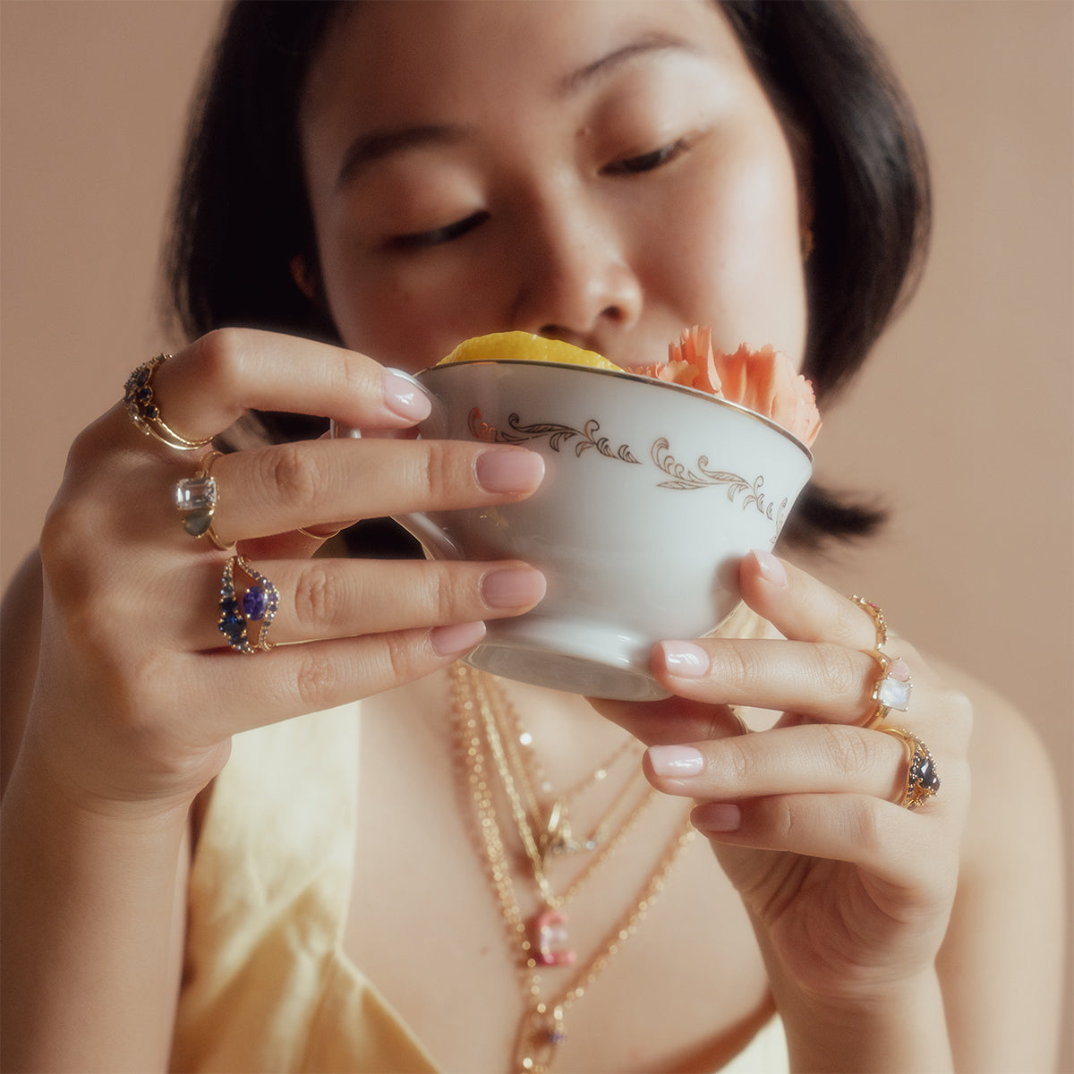 A woman wearing gold jewelry and the True Colors Classic Ombre Birthstone Ring holds a decorative teacup filled with colorful flowers, gazing at it closely against a soft beige background.