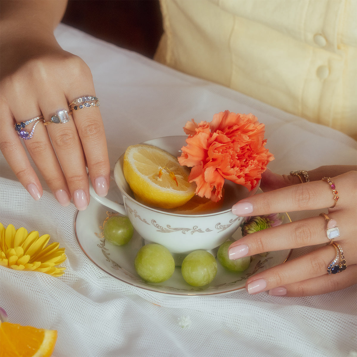 A close-up of hands with pastel manicured nails and multiple rings, including the True Colors Classic Ombre Birthstone Ring, holding a teacup with lemon and orange carnation. The saucer holds green grapes, a yellow flower, on white cloth.