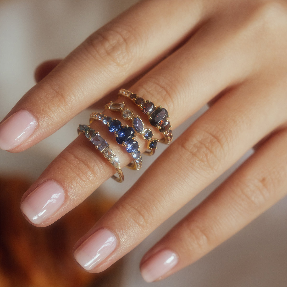 A hand with neatly manicured nails wears multiple 14k gold plated rings, including the Shades of You Ombre Birthstone Ring featuring blue, purple, and clear gemstones stacked on the middle and ring fingers, against a softly blurred background.