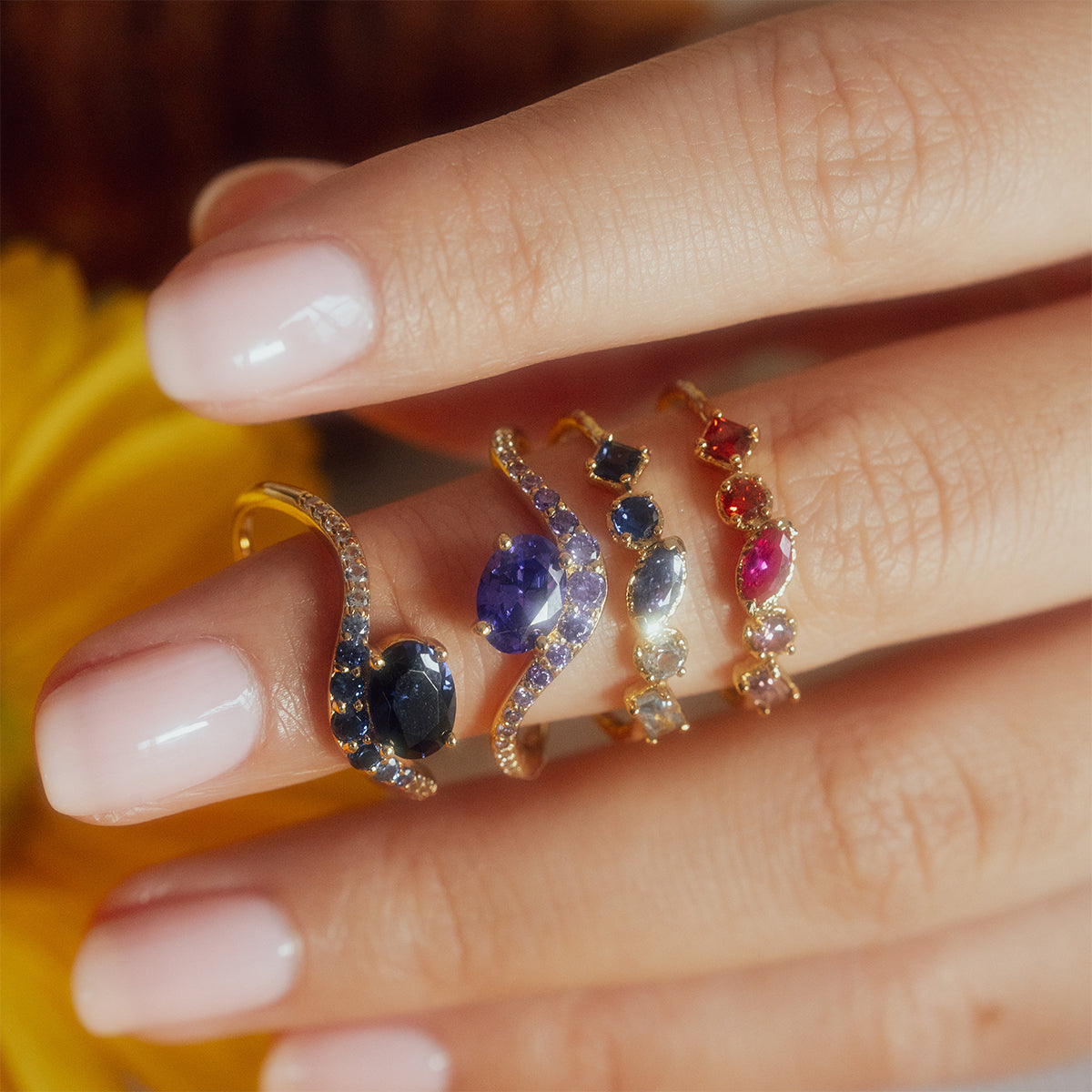 A close-up of a hand with light pink nails wearing four Shades of You Ombre Birthstone Rings, each 14k gold plated and set with colorful blue, purple, and red gemstones, with a yellow flower in the background.