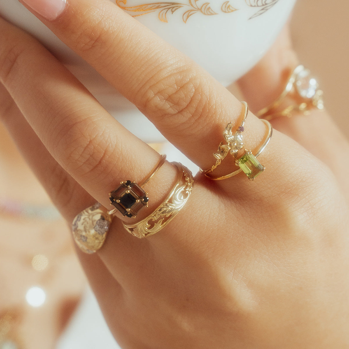 A close-up of manicured fingers wearing the Solid Gold Essential Baguette Birthstone Ring with natural gemstones, holding a white teacup decorated with a gold pattern.
