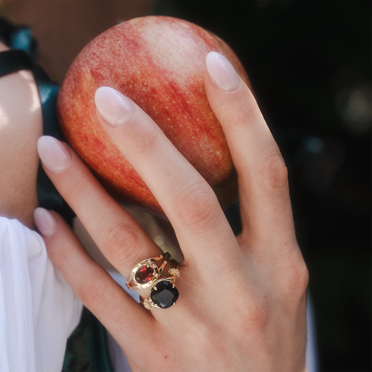A hand with long, pale pink nails holds a red apple, adorned with the Past Life Black Spinel Statement Ring and a gold ring with a red gemstone. The person is wearing a white sleeve.