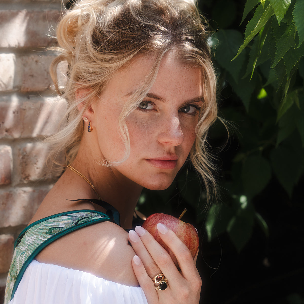 A young woman with wavy blonde hair stands by a brick wall with green leaves, holding a red apple and wearing a white off-shoulder top paired with Sapphire Soliloquy Huggie Hoops.