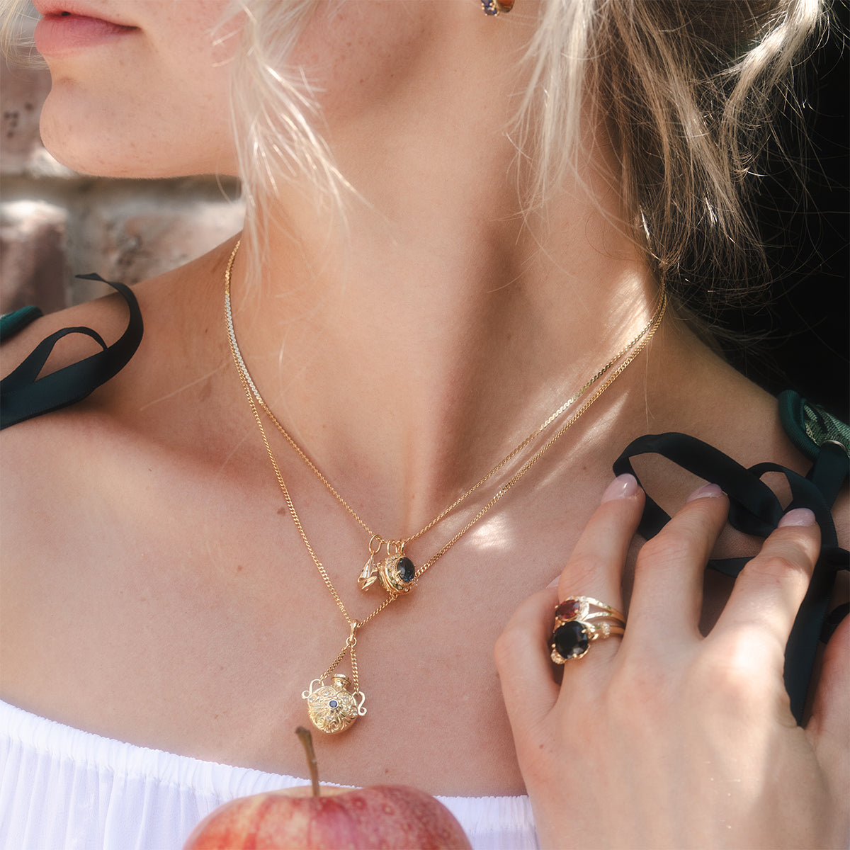 A woman with light hair holds a red apple, wearing layered gold gemstone necklaces and the Past Life Black Spinel Statement Ring, paired with an off-shoulder white top featuring dark green ribbons.