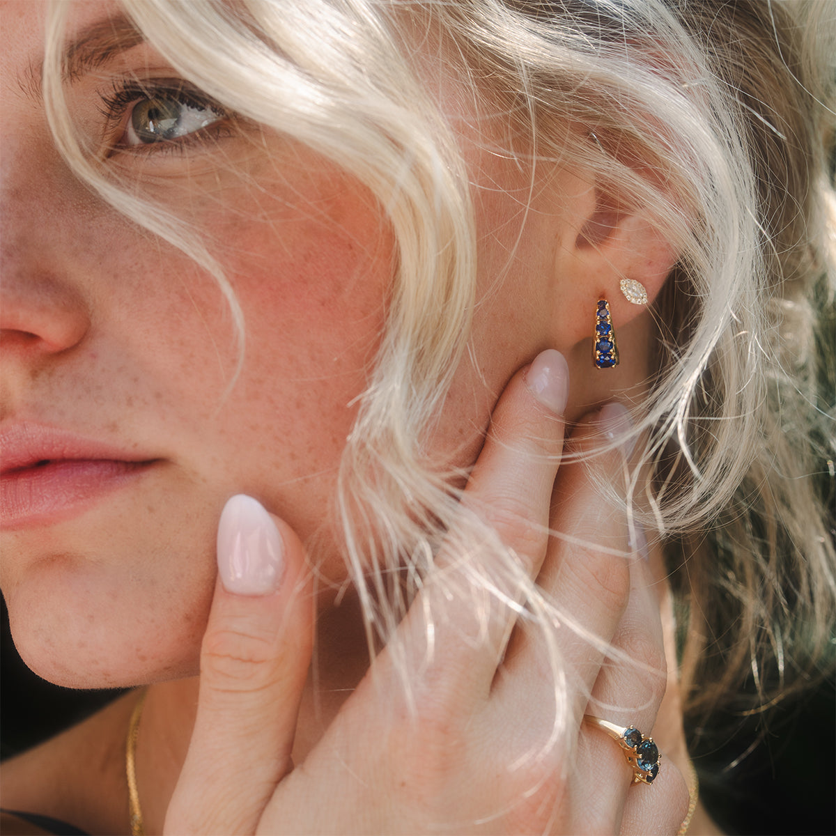 Close-up of a woman with wavy blonde hair wearing the Sapphire Soliloquy Huggie Hoops—vintage-inspired gold earrings set with blue sapphires—and a matching 14k gold vermeil ring, elegantly highlighting these stunning pieces.