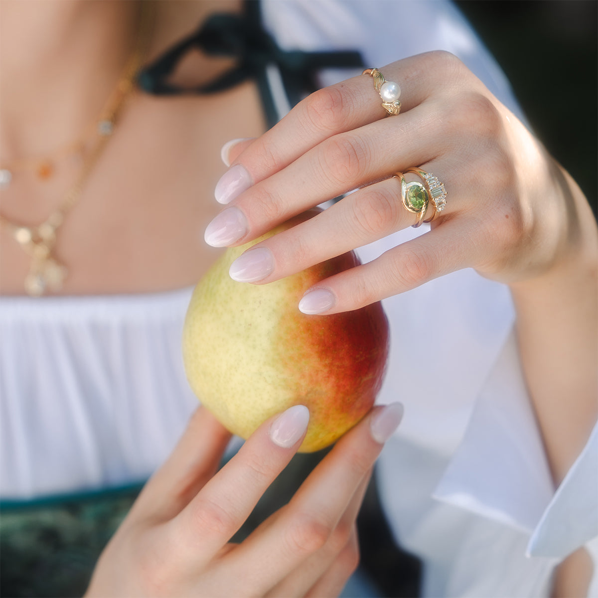 A person with manicured nails holds a red and yellow apple with both hands, wearing a white blouse, black ribbon, gold necklace, and the Peridot Poetry Statement Ring.