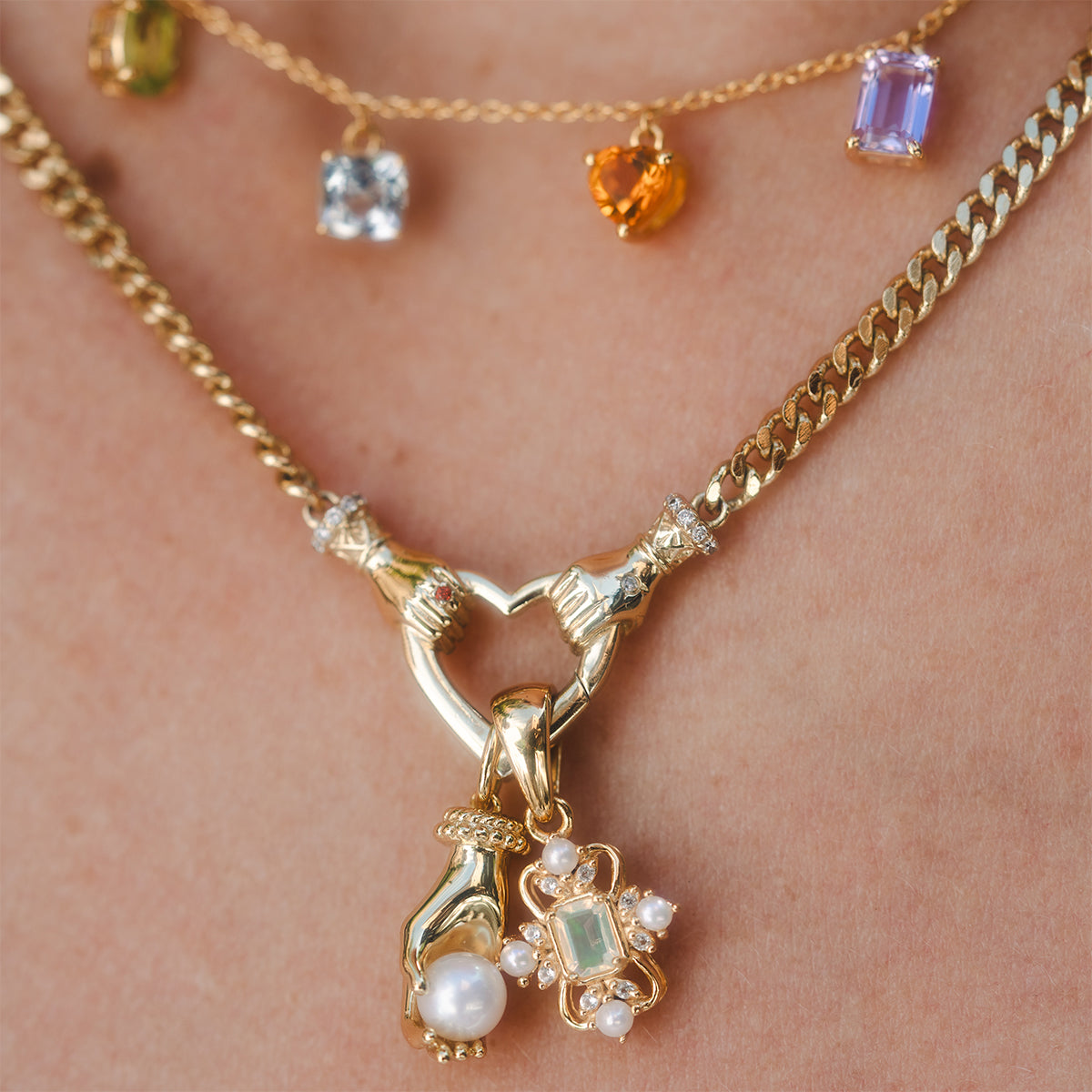 Close-up of a person wearing layered gold necklaces, featuring the Hand Over Heart Charm Holder Necklace, a pendant with pearls and gemstones, and another necklace adorned with colorful gemstone charms.