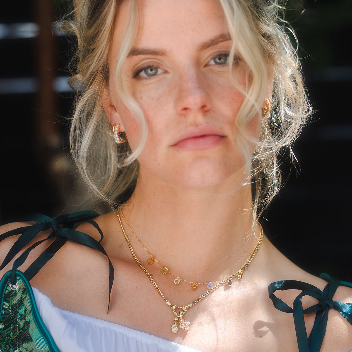 A woman with loose blonde curls wears Pearl Keepsake Huggie Hoops and layered gold charm necklaces, paired with a white off-shoulder top featuring dark green ribbon straps, standing outdoors in soft natural light.
