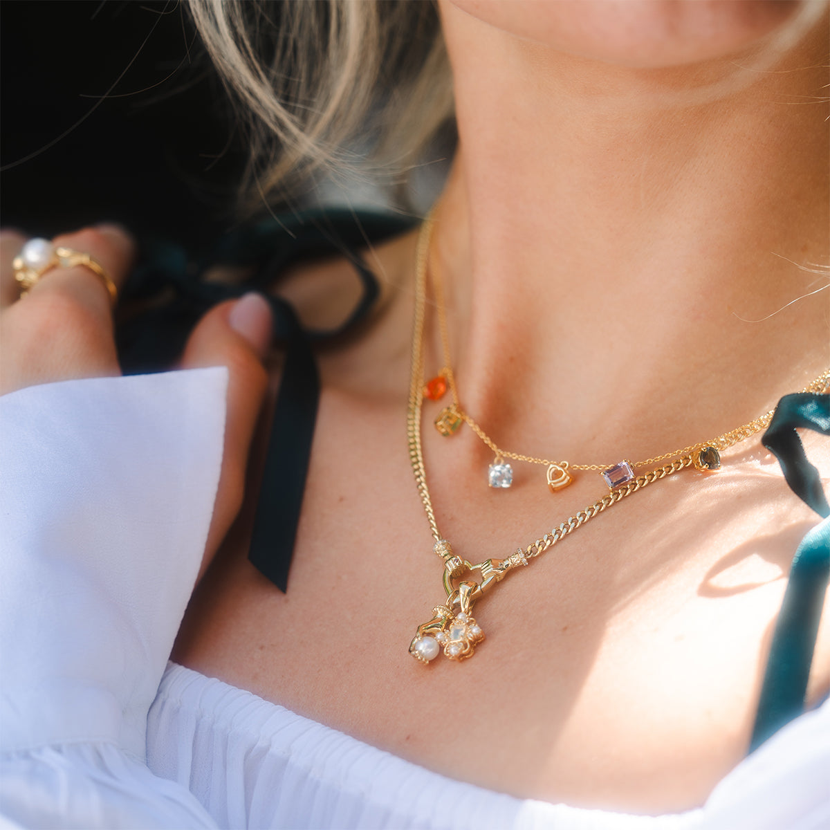 A woman wears the Hand Over Heart Charm Holder Necklace in 14k gold vermeil with gemstone charms and pearls, layered with other gold pieces and a ring. She pairs it with a white off-shoulder top featuring black ribbon straps.