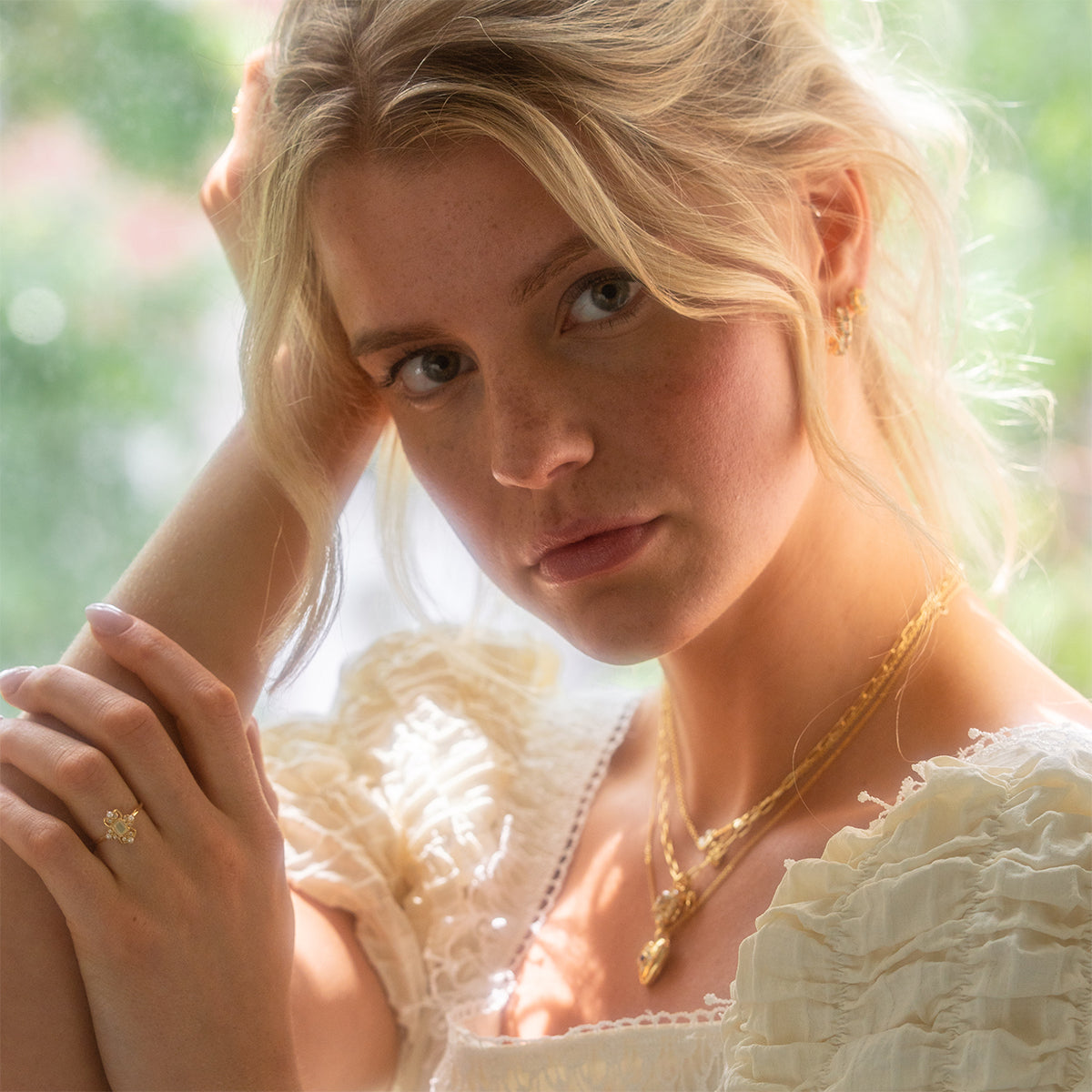 A young woman with blonde hair in loose waves wears a ruffled white blouse and 14k gold vermeil jewelry, including necklaces, earrings, and the Crown Jewel Opal and Pearl Ring, gazing softly at the camera in natural light.