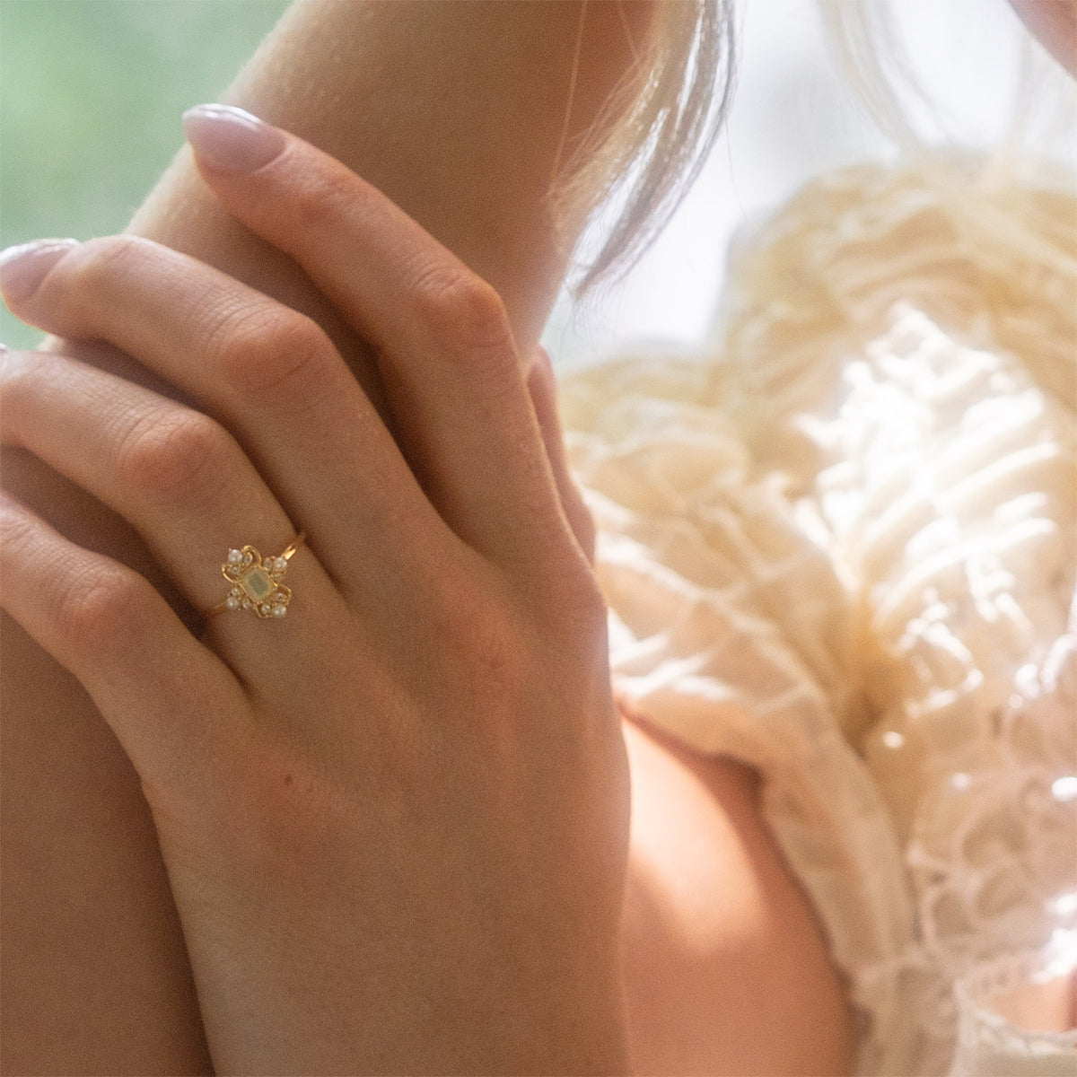 A close-up of a womans manicured hand wearing the Crown Jewel Opal and Pearl Ring in 14k gold vermeil with a floral design, styled with a ruffled cream blouse and bathed in soft natural light for vintage-inspired charm.