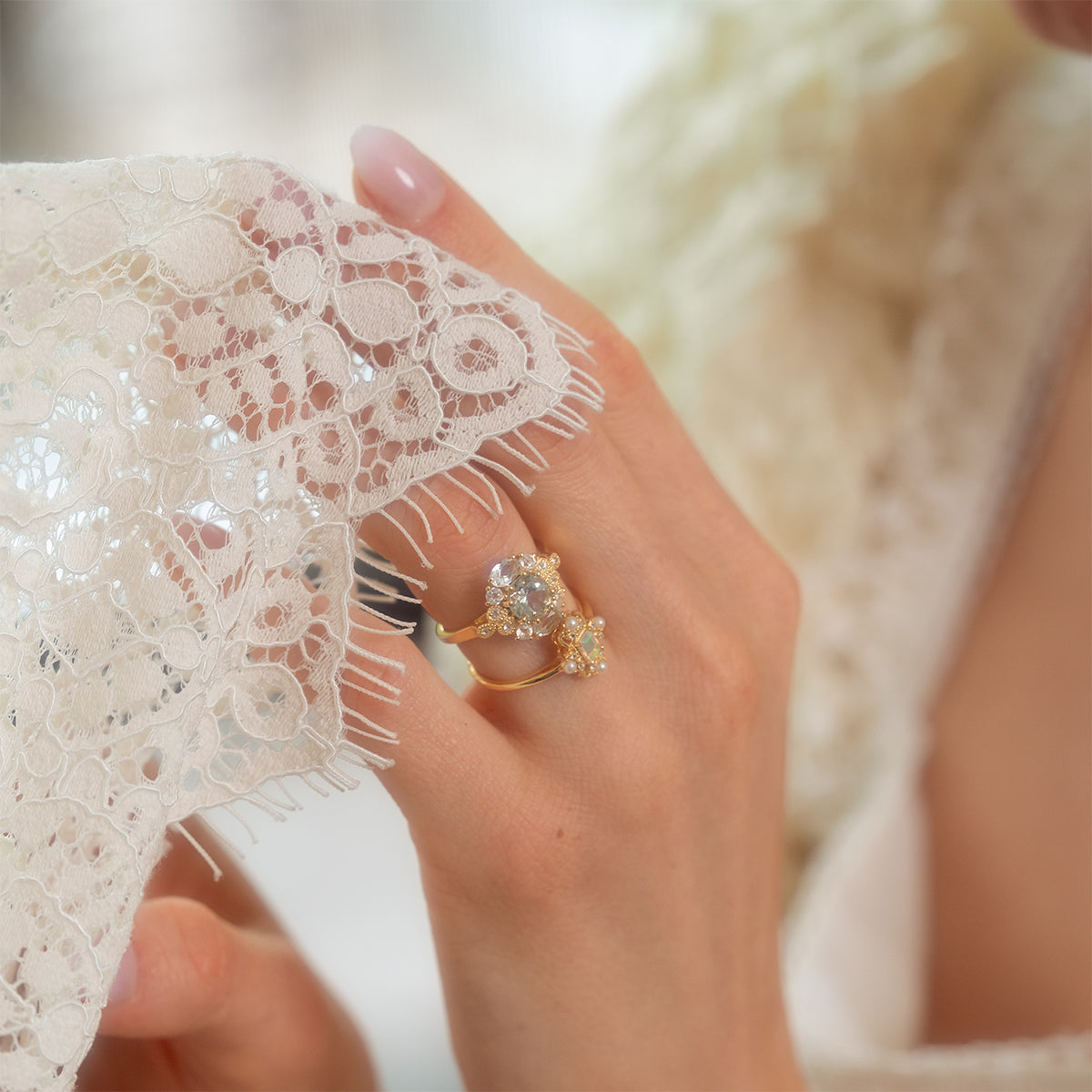 A close-up of a hand holding delicate white lace fabric, wearing the Royal Gardens Green Amethyst Cocktail Ring—gold with sparkling multicolored gemstones, including green amethyst. The scene exudes a soft and elegant feel.