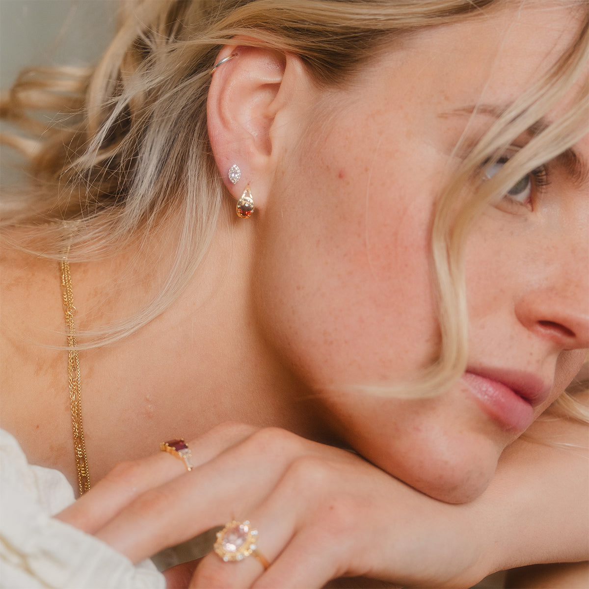 A blonde woman leans her chin on her hand, showcasing gold jewelry, including the Gilded Garnet Huggie Hoops, a stud earring, a gold ring with white topaz, and a delicate necklace.