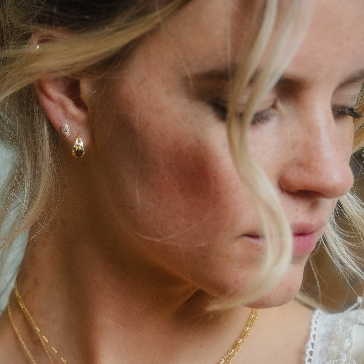 A woman with blonde hair wears a white dress, a gold necklace, and Gilded Garnet Huggie Hoops.