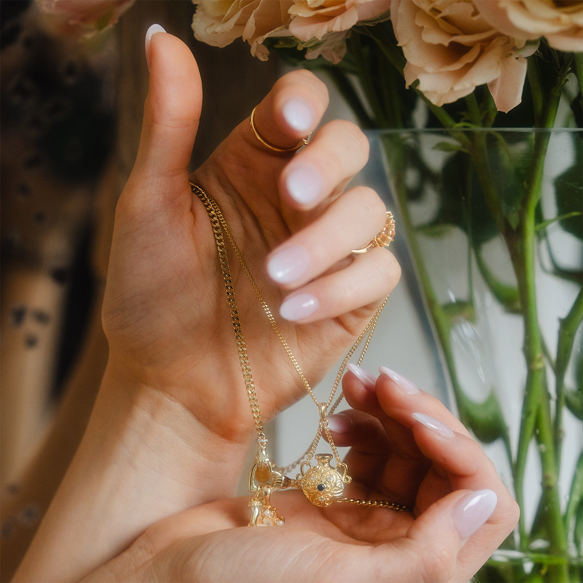A close-up of manicured hands holding the Juliet Vial Pendant Necklace, a vintage-inspired gold jewelry piece, in front of a glass vase filled with light pink flowers.