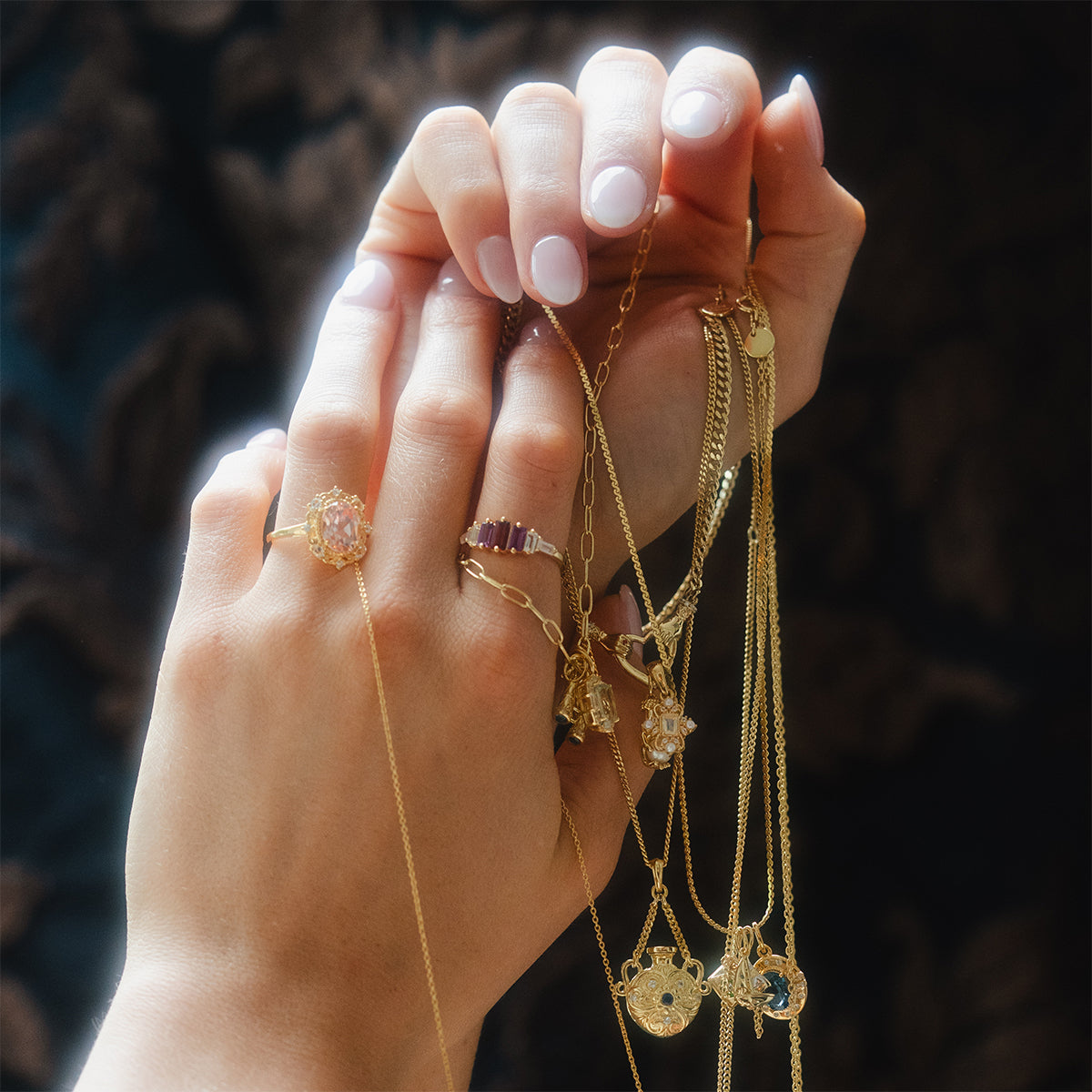 Hands with light pink nail polish hold gold necklaces featuring the Vintage Inspired Trinket Charm, paired with gemstone gold rings, against a blurred dark background.