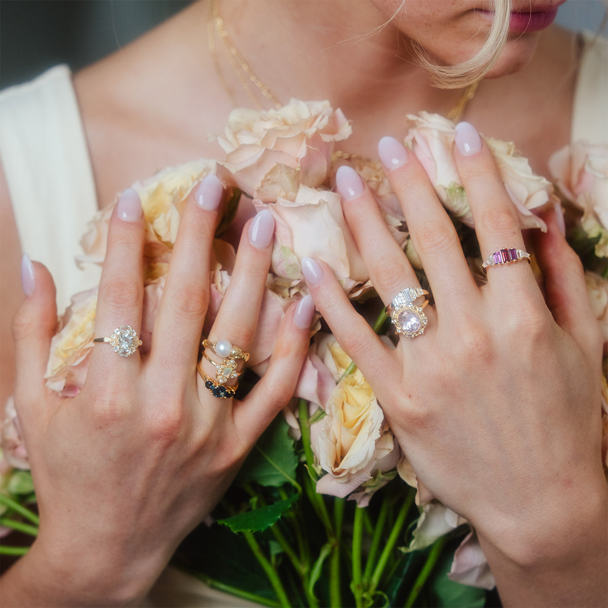 A woman in a light-colored dress holds pale pink roses, showcasing ornate rings—including the Royal Gardens Green Amethyst Cocktail Ring—on her manicured fingers. Only her hands, part of her face, and dress are visible.