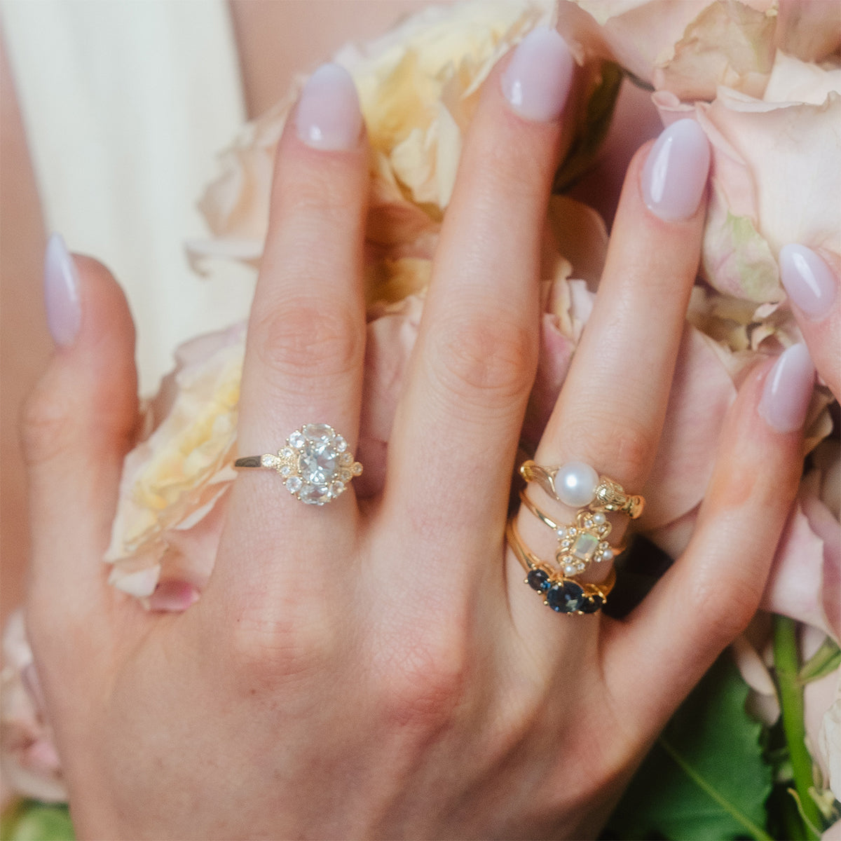A hand with pastel pink manicured nails, adorned with several gold rings including the Royal Gardens Green Amethyst Cocktail Ring, gently holds pale pink roses.