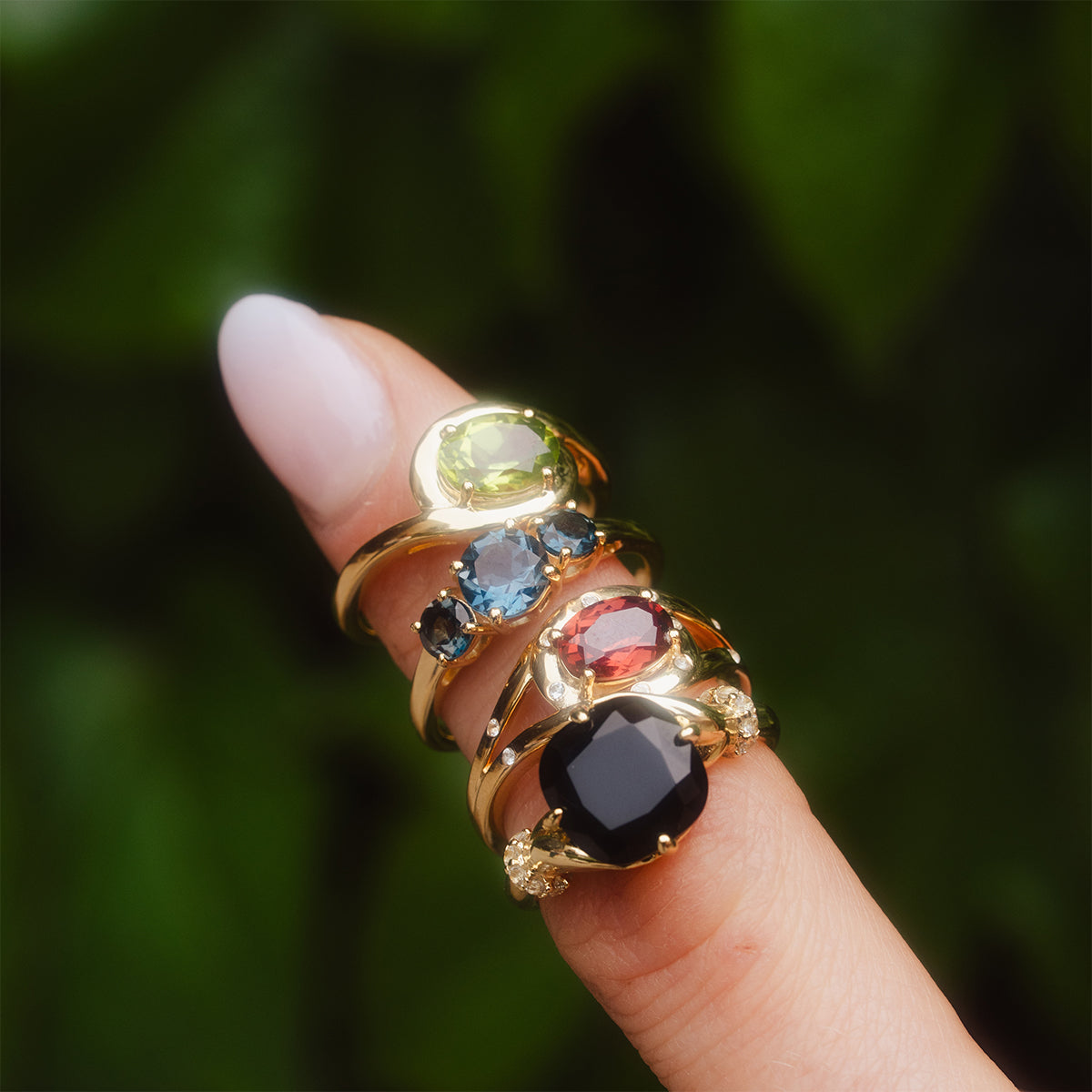 A hand with a pale pink manicure showcases the Past Life Black Spinel Statement Ring alongside vintage-inspired gold rings with large light green, blue, and red gemstones, set against a blurred green background.