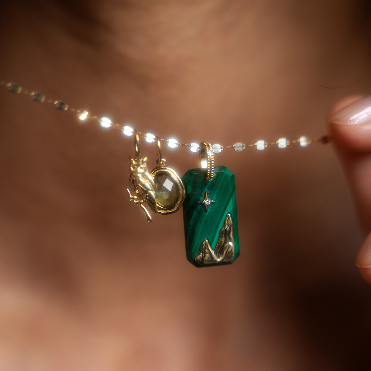 A close-up of a hand holding a Solid Gold Amulet Charm necklace, featuring three gold charms: a small figure, an oval gem, and a green pendant with gold mountain designs and an embedded star—a local eclectic exclusive.