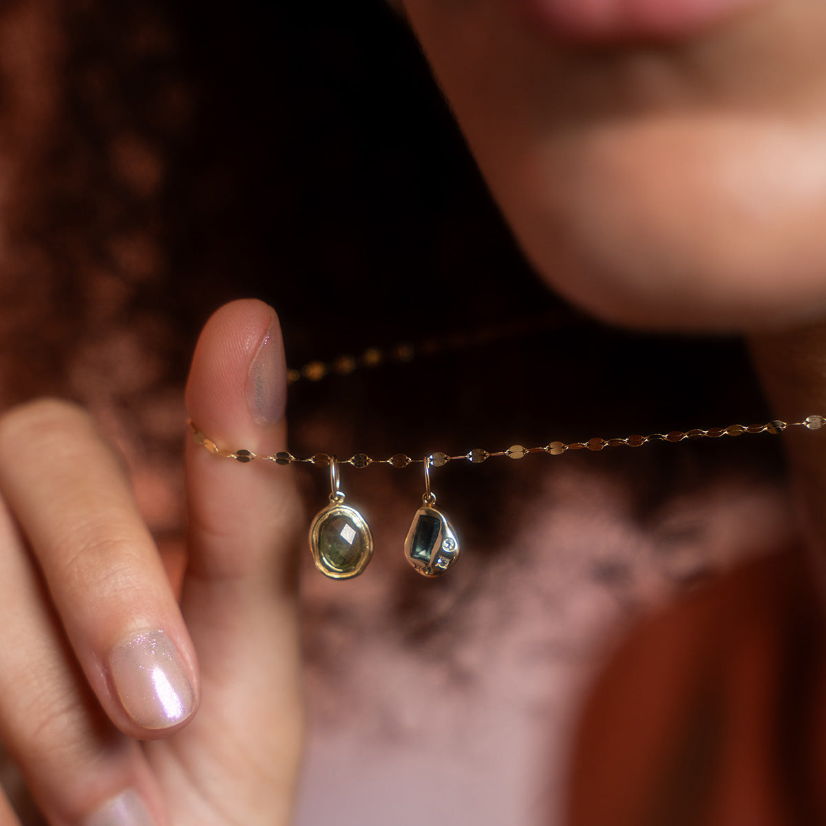 A person with curly hair wears a local eclectic exclusive Solid Gold Amulet Charm necklace, featuring two pendants—an oval gemstone and a small skull-shaped solid gold charm. Only their lips, part of their face, and hand are seen in the image.