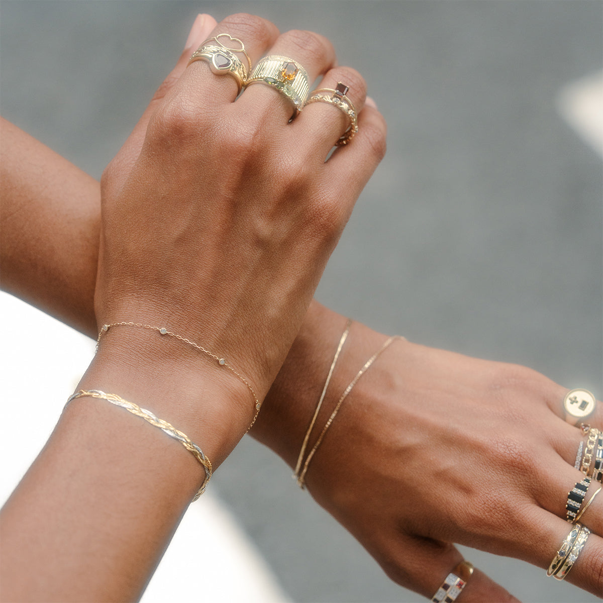 Two hands wearing multiple gold rings and the Solid Gold Everyday Opal Bracelet, featuring delicate textures and subtle opal gemstone accents, are shown against a blurred neutral background.