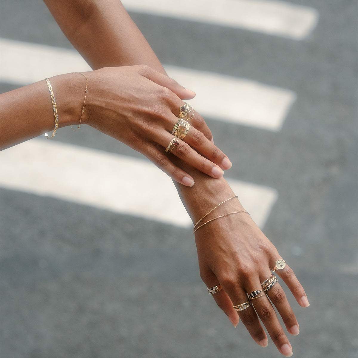 Two hands adorned with the Solid Gold Black Agate Crown Ring and thin gold bracelets, gracefully held together, with a blurred street and crosswalk lines in the background.
