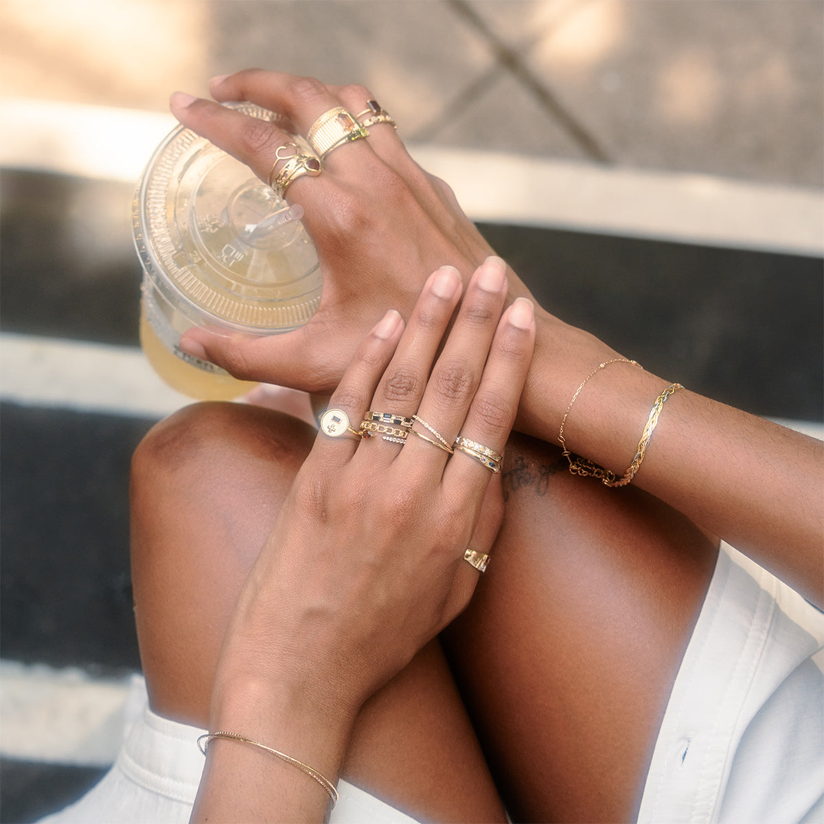 Wearing the Solid Gold Delicate White Sapphire Stacking Band and bracelets, a person in a white outfit holds an iced drink with crossed legs outdoors, sunlight and shadows dancing on the pavement.