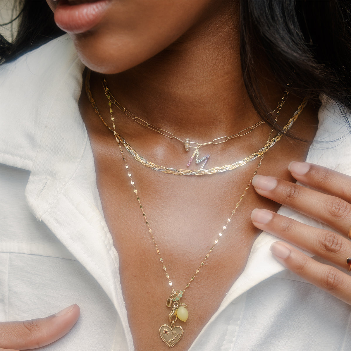 A woman in a white shirt displays layered solid gold necklaces, featuring the Solid Gold Mixed Metal Braided Necklace, a heart pendant, and another necklace with a colorful M charm. She softly touches her collar.