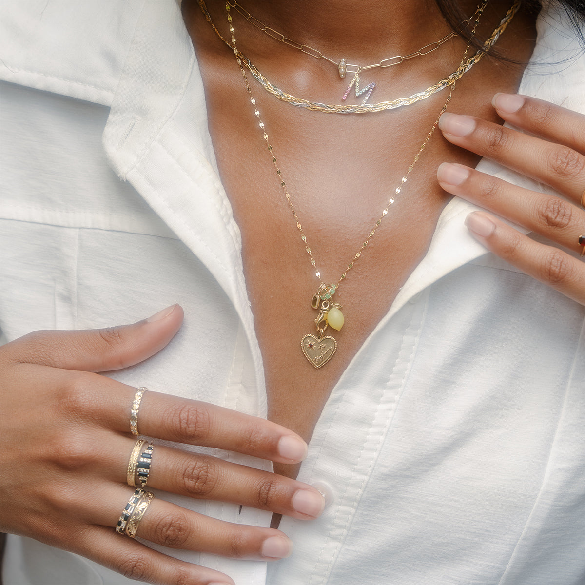 A woman in a white collared shirt with an open neckline wears layered gold necklaces—a heart locket and an M initial charm—while displaying several rings, including the Solid Gold Black Agate Crown Ring.