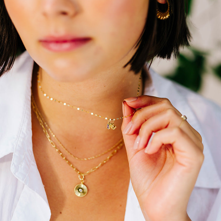 A woman with short dark hair wears layered gold necklaces, featuring the Solid Gold Birthstone Baguette Charm and an initial charm chain, highlighting personalized jewelry as she gently holds one necklace. She is dressed in a white collared shirt.