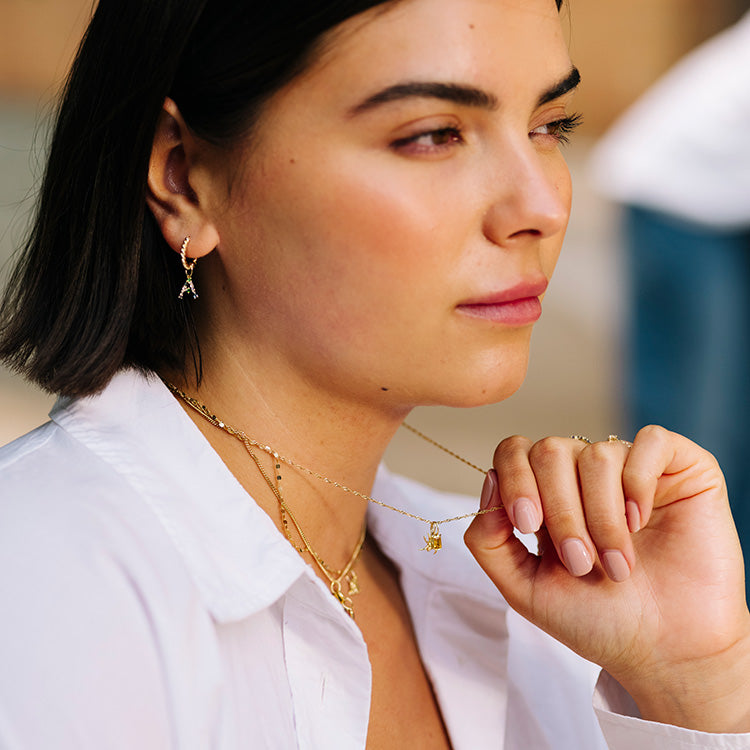 A woman with short dark hair in a white blouse gazes to the side, wearing gold layered necklaces and matching earrings adorned with Eiffel Tower charms and a sparkling Solid Gold Birthstone Baguette Charm.