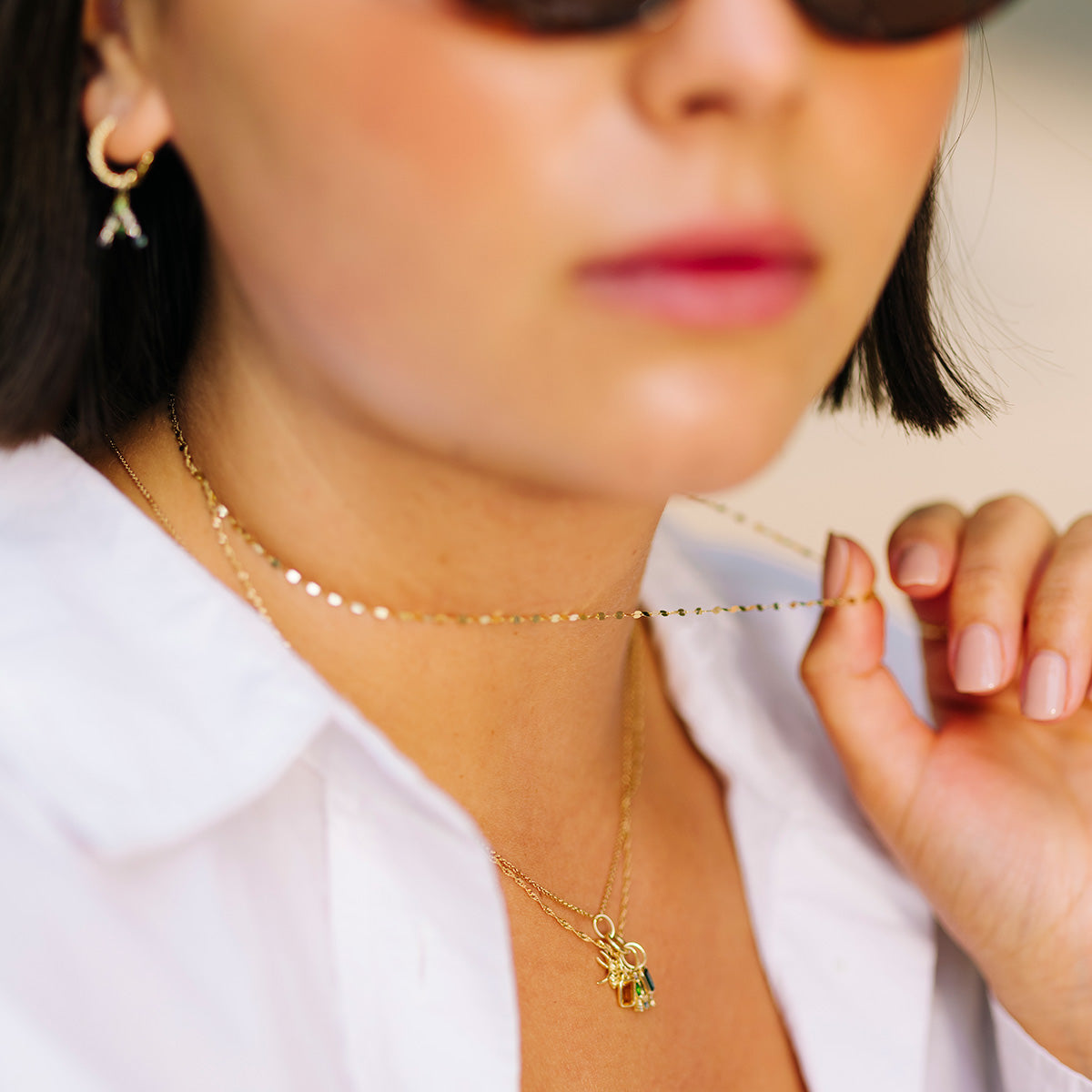 A woman in sunglasses and a white shirt holds the Solid Gold Sparkle Link Chain with two pendants. She also wears small gold hoop earrings and has neatly manicured nails, with her face partially out of focus.