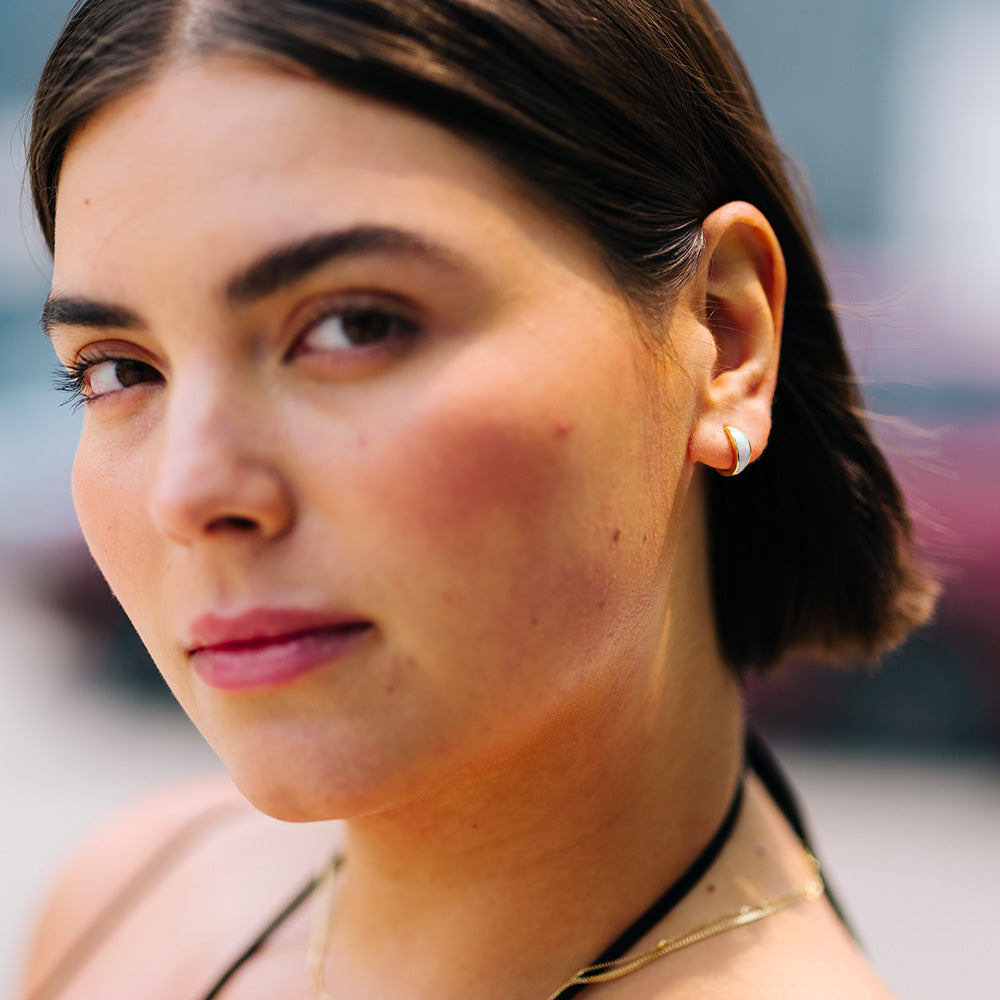 A woman with short brown hair wears Solid Gold Chunky Opal Inlay Huggie Hoops and a black halter top. With natural makeup, she looks confidently at the camera, set against a soft background.