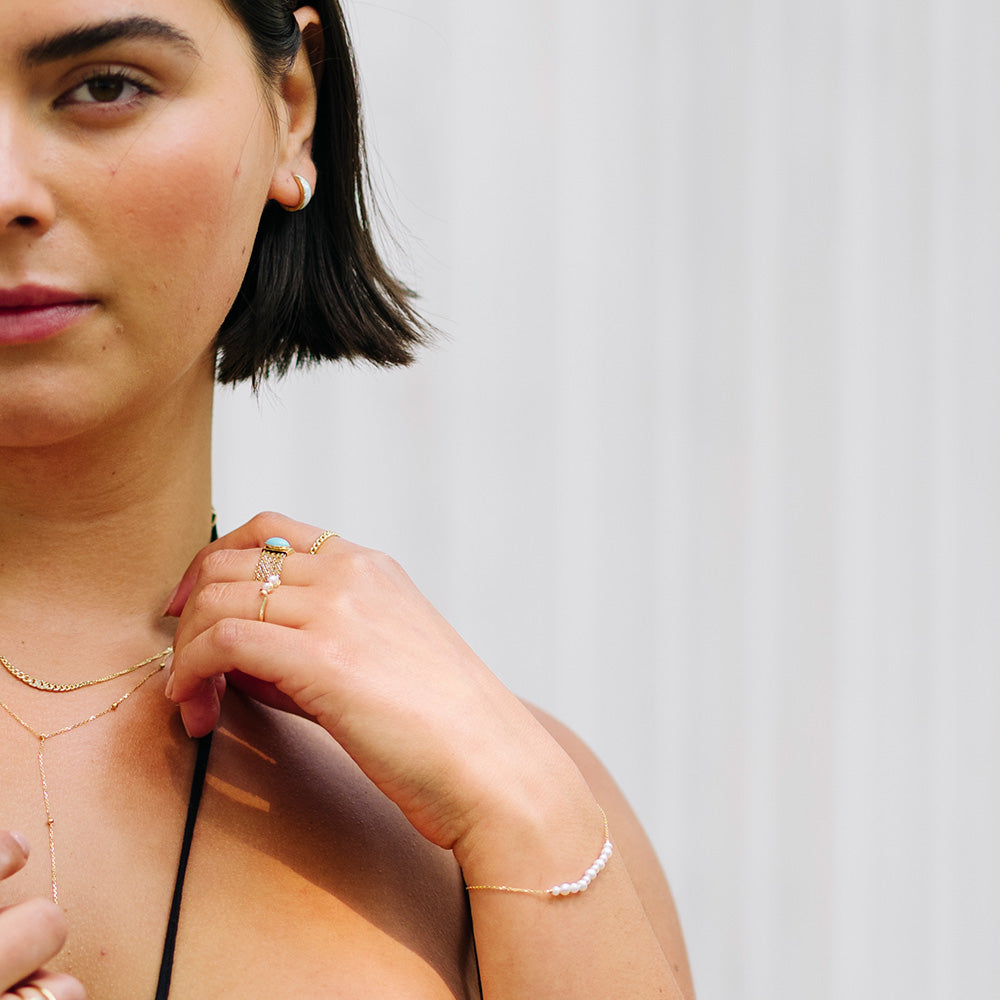 A woman with short dark hair, wearing gold rings, layered necklaces, and the Solid Gold Pearl Strand Bracelet with freshwater pearls, is shown from the shoulders up against a light striped background with a neutral expression.