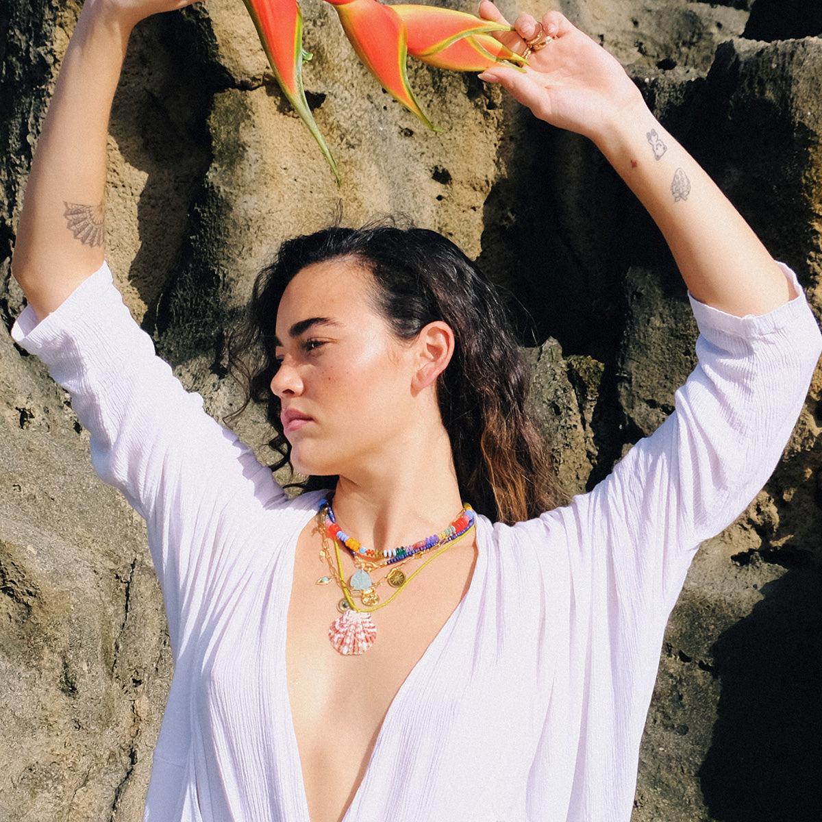 A person in a white, deep V-neck top stands against rocks, holding an orange flower above their head. They wear the Rainbow Candy Necklace layered with other pieces and have small tattoos visible on their arms.